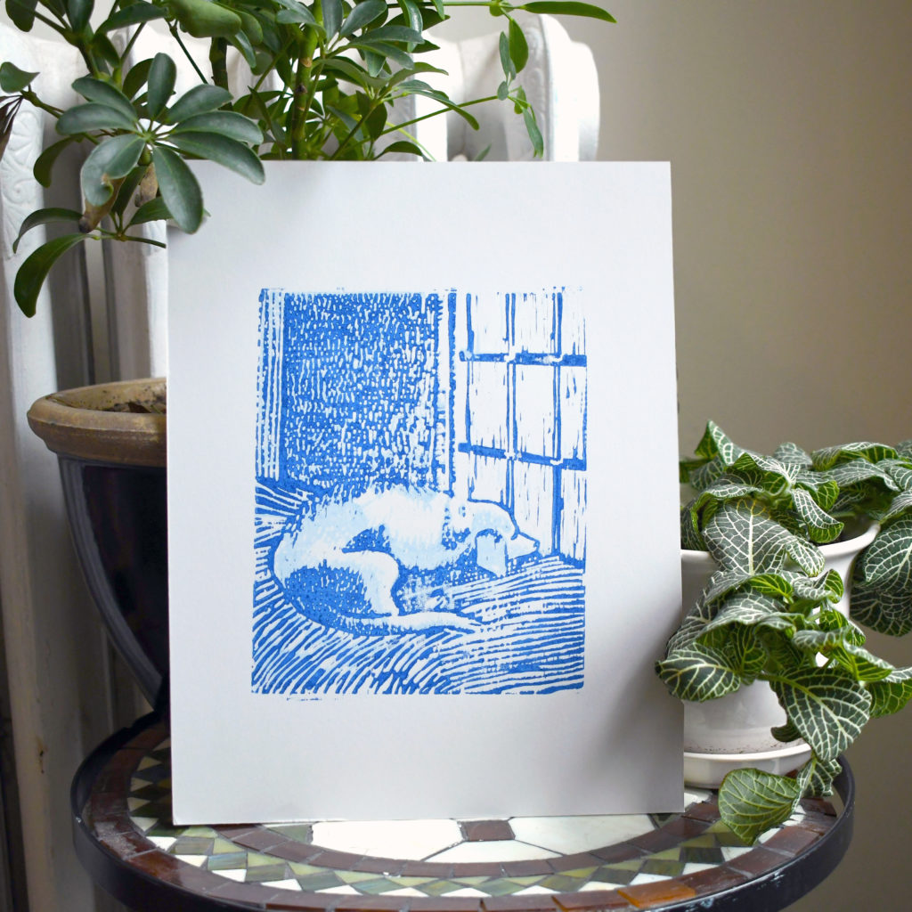 blue woodcut art print in a dog lying on a beg looking out the window