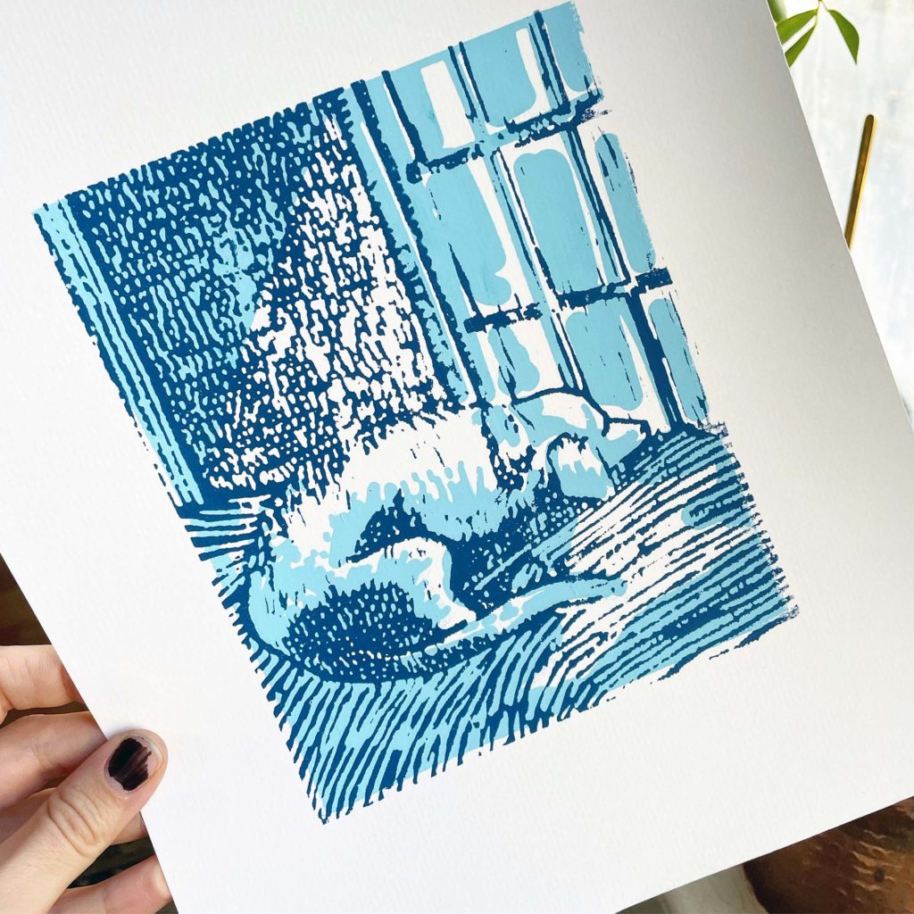 A hand holds up a dark blue and light blue print of a dog sleeping by a window.